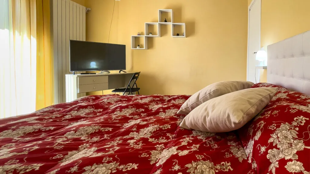 Camera 99 Cannelle: letto matrimoniale con copriletto rosso a fiori, televisore a parete e mensole bianche
